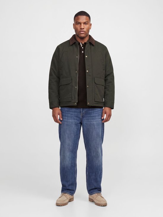 Model draagt een donkergroene jas over een bruine poloshirt, blauwe jeans en beige schoenen.