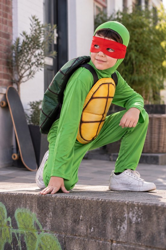 Kind in groen Ninja Schildpad kostuum met rood masker, gehurkt op een muur met een skateboard op de achtergrond.