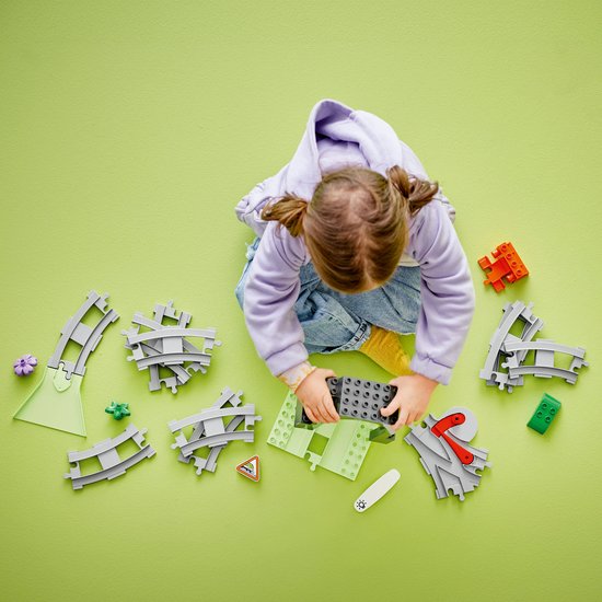 Een meisje met staartjes speelt met Duplo treinspoorstukken, verspreid op een groen kleed.