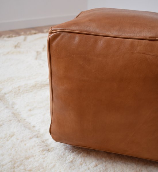 Caramelbruine vierkante poef van leer, op een wit vloerkleed.