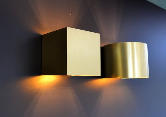 Twee wandlampen, één vierkant mat goud en één cilindervormig messing, verlichten de muur.