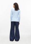 Model van achteren met een lichtblauw geribbeld shirt, donkere jeans met wijde pijpen en krullend haar.