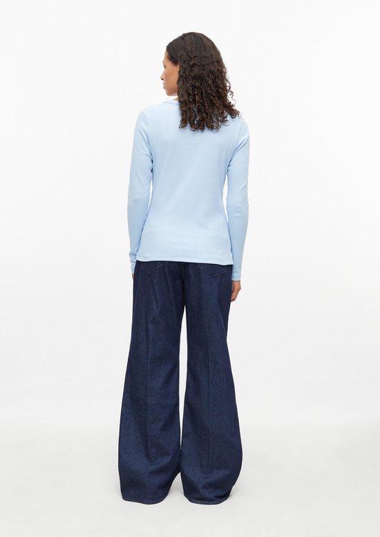 Model van achteren met een lichtblauw geribbeld shirt, donkere jeans met wijde pijpen en krullend haar.