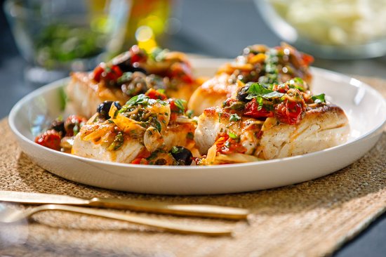 Gegrilde vis met tomaten, olijven en basilicum op een wit bord.