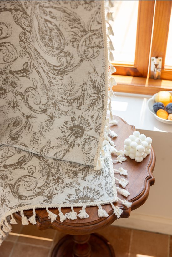 Crèmekleurige gordijnrand met bruine barokprint en decoratieve, witte franjes.