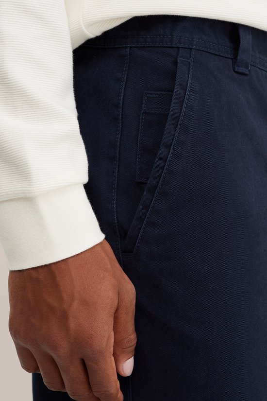 Detail van de zak van een donkerblauwe chino, gedragen door een model.