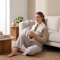 Een vrouw met een beige trui en grijze broek zit op de vloer bij een bank en houdt een mok vast, met een nekwarmer om.