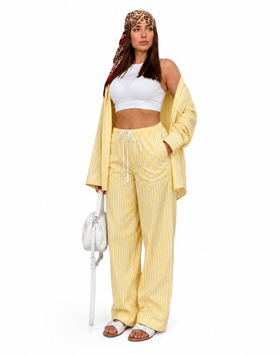 Vrouw in gele gestreepte pyjamaset, witte crop top, sjaal met luipaardprint en witte tas.