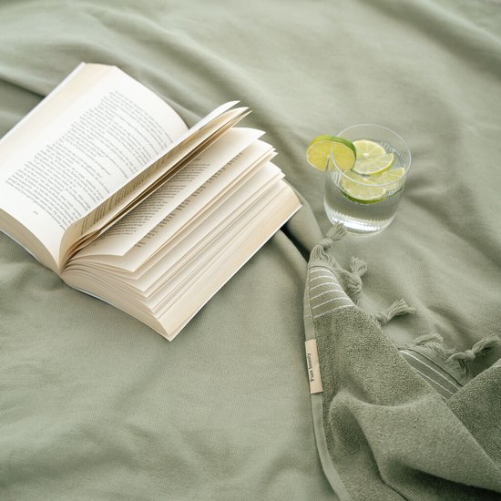 Een open boek, glas water met limoen en een groene hamamdoek met franjes op een groen bed.