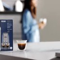 Close-up van een doos De'Longhi EcoDecalk met een glas espresso op de voorgrond.