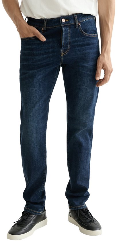 Man met de handen in de zakken van een donkerblauwe jeans. Hij draagt een wit T-shirt en zwarte sneakers.