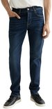 Man met de handen in de zakken van een donkerblauwe jeans. Hij draagt een wit T-shirt en zwarte sneakers.