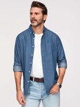 Man met baard, draagt een blauw overhemd met witte stipjes, een wit t-shirt, spijkerbroek en bruine riem.