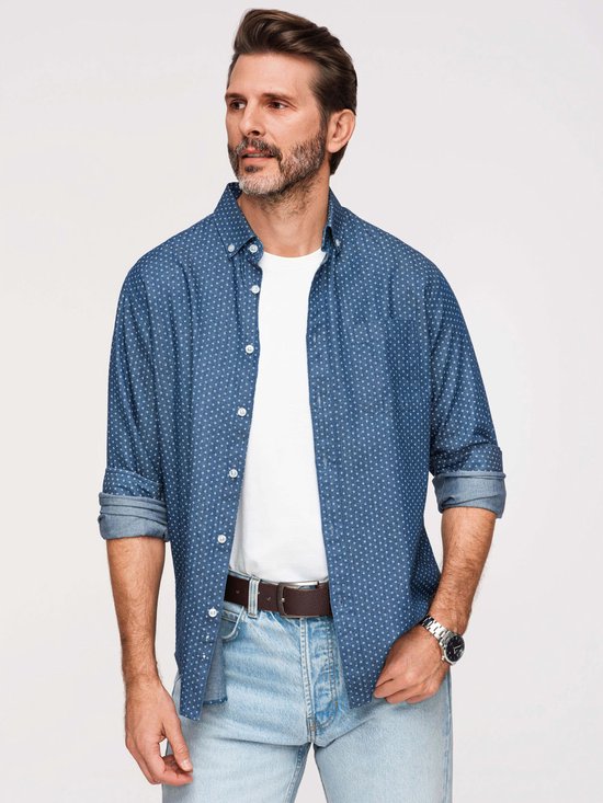 Man met baard, draagt een blauw overhemd met witte stipjes, een wit t-shirt, spijkerbroek en bruine riem.
