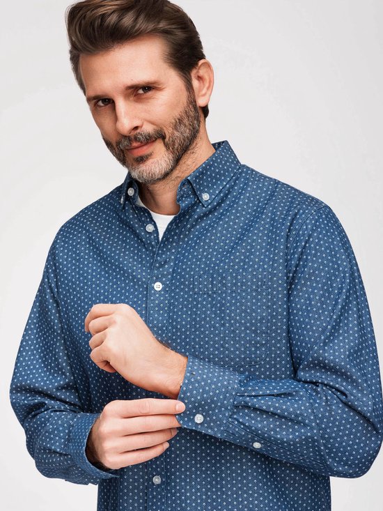 Man met baard, draagt een blauw overhemd met witte stipjes, wit t-shirt, spijkerbroek, stopt mouw op.