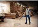 Man in beige vest blaast houtstof weg van de vloer met blauwe Bosch bladblazer in een werkplaats.