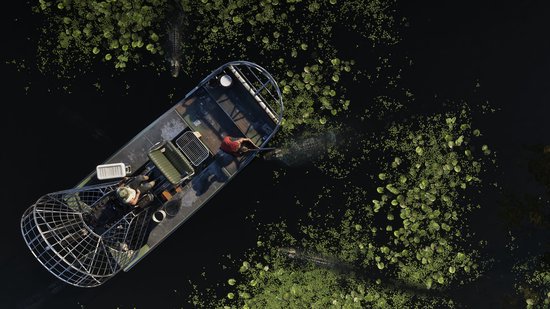 Luchtfoto van een airboat met twee personen die op het water varen, omringd door waterlelies en enkele alligators.