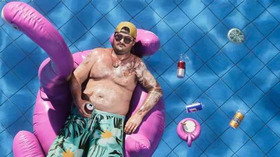 Man met een pet en zwembroek, drijvend op een roze opblaasbare flamingo in een zwembad, met blikjes drankjes.