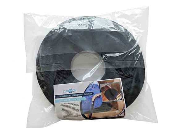 Zwart donutkussen van schuim met een gat in het midden, verpakt in plastic. Een label toont een persoon die op een stoel zit.