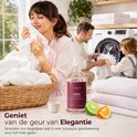 Vrouw ruikt aan schone was met een Steamstory Wasparfum Love fles, sinaasappel, bergamot en witte bloemen. Gezin wast op de achtergrond.