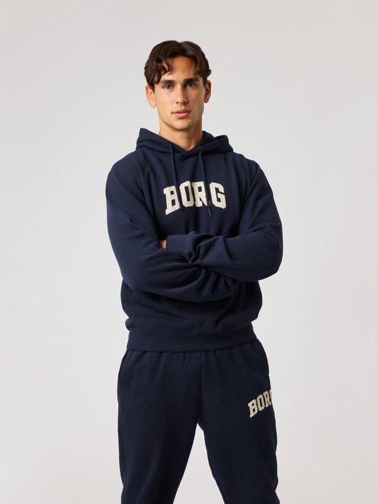 Man met armen over elkaar in een donkerblauwe hoodie met crème 'BORG' opdruk en bijpassende broek.
