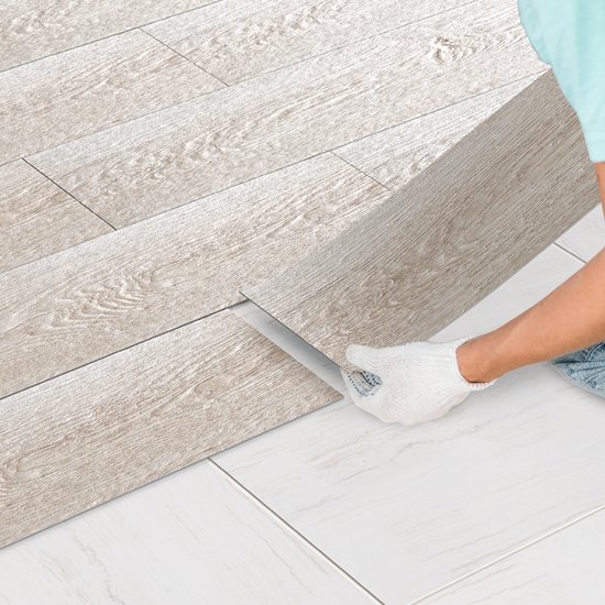 Handen in werkhandschoenen leggen zelfklevende eikenhouten pvc-planken op een witte tegelvloer.