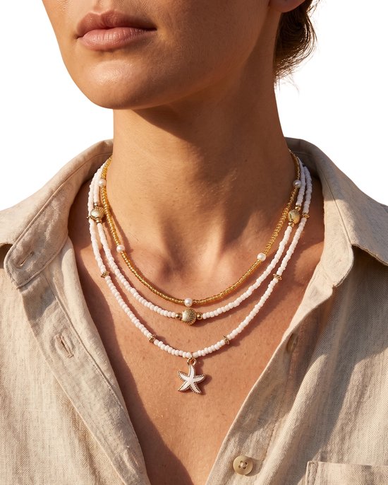Model draagt drie gouden kettingen met witte en gouden kralen, schelp, parel en sterhanger.