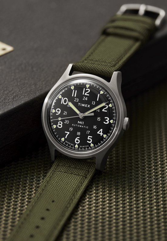 Zilverkleurig Timex MK1 Automatic horloge met zwarte wijzerplaat en groene stoffen band, naast een donkere doos.