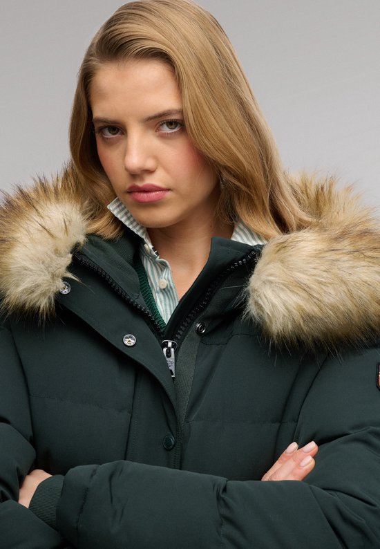 Model met een donkergroene, gewatteerde bomberjack met bruine bontkraag. Onder de jas draagt ze een gestreepte blouse en groen vest.