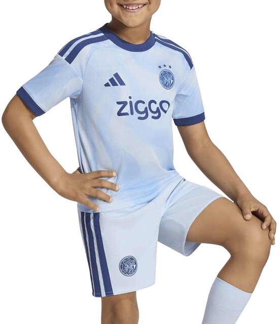 Jongen in lichtblauw Ajax-shirt met donkerblauwe accenten en short. Adidas-logo en 'ziggo'-opdruk.