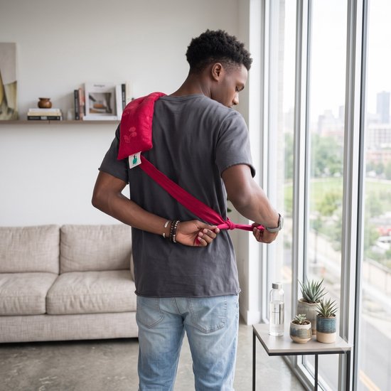 Man met grijs T-shirt knoopt achter zijn rug een rood warmtekussen met linten vast.