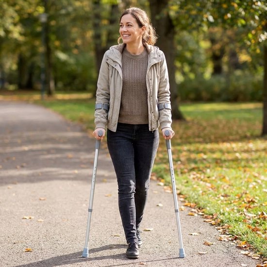 Een lachende vrouw loopt op een pad in een park met twee grijze elleboogkrukken.