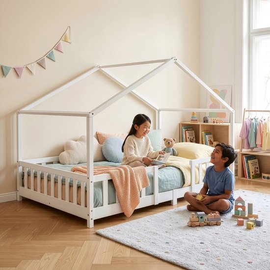 Deux enfants sur un lit maison blanc avec barreaux, avec coussins et couvertures.