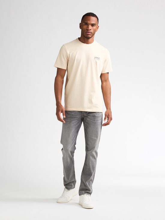 Man staat recht naar voren, draagt een crème T-shirt met opdruk en grijze jeans.