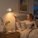 Witte, dimbare wandlamp boven een bed met een vrouw die een boek leest en de lamp bedient met afstandsbediening.