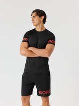 Man in zwart sportshirt met rode Björn Borg letters op de mouwen, met gekruiste armen. Zwarte sportshort met rode letters.