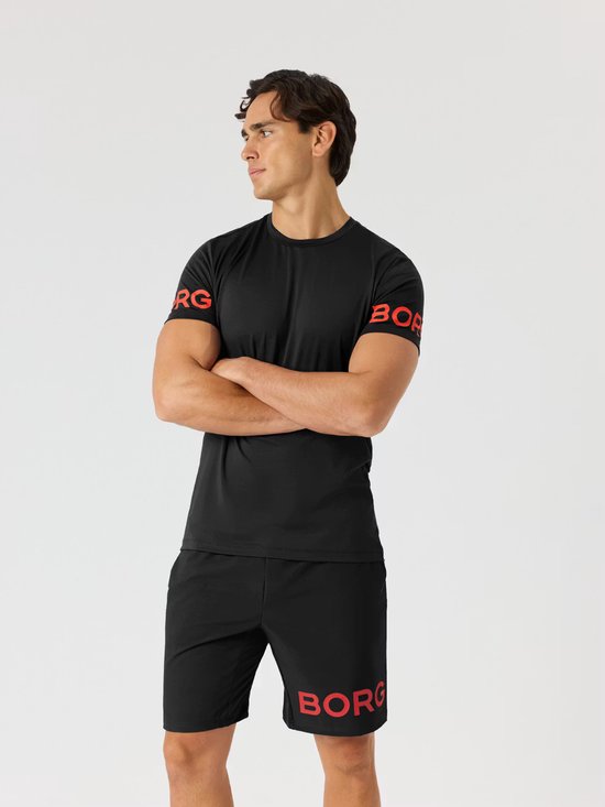 Man in zwart sportshirt met rode Björn Borg letters op de mouwen, met gekruiste armen. Zwarte sportshort met rode letters.