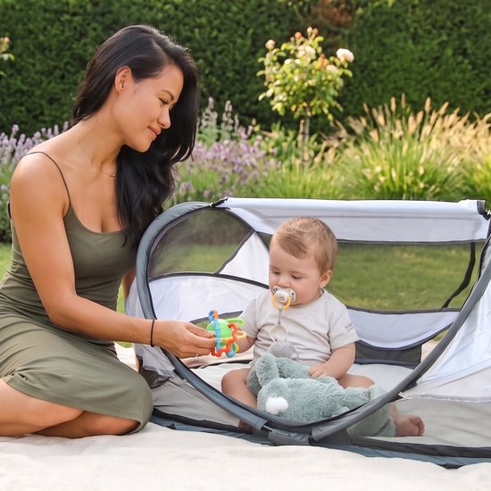 Moeder met baby in grijs-wit Deryan campingbedje in de tuin op een kleed.