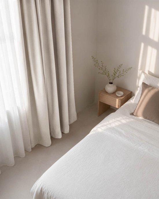 Licht beige verduisterend gordijn met ringen, deels voor een wit raam, naast een houten nachtkastje met een vaas.