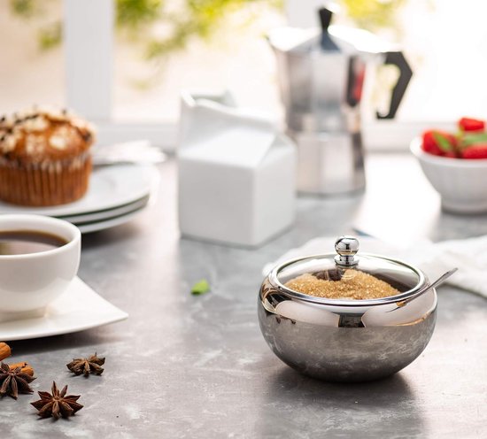 Zilverkleurige suikerpot met deksel, gevuld met bruine suiker, naast een kop koffie en een muffin.