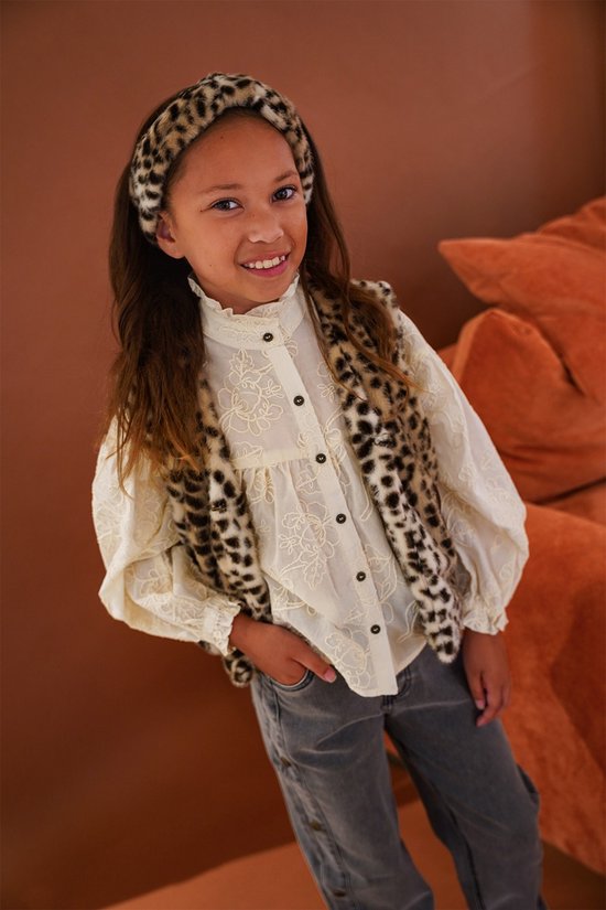 Meisje met luipaardprint bodywarmer, gebloemd wit blousje met pofmouwen, grijze jeans en haarband met luipaardprint.