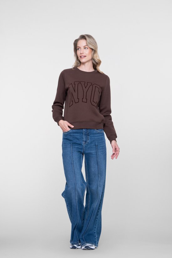 Model met blonde krullen in een donkerbruine sweater met 'NYC' opdruk en een wijde blauwe jeans, staand in een studio.
