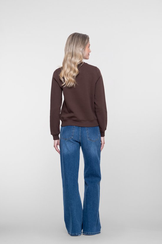 Vrouw met blonde krullen, staand in een donkerbruine sweater met 'NYC' opdruk en een blauwe jeans met wijde pijpen.