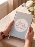 Man houdt grijs boek met de tekst "Lieve papa, ik ben zo nieuwsgierig..." vast, met bloemen op tafel.