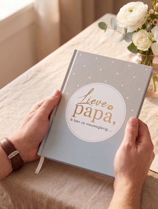 Man houdt grijs boek met de tekst "Lieve papa, ik ben zo nieuwsgierig..." vast, met bloemen op tafel.