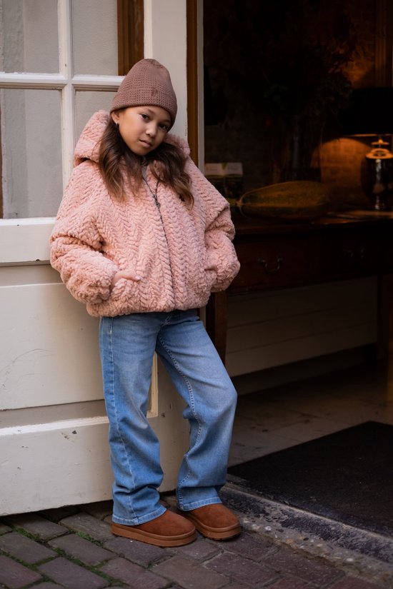 Meisje met bruine muts staat in roze teddy jas en spijkerbroek.