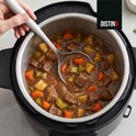 Binnenkant van een zwarte DistinQ multicooker gevuld met stoofvlees, aardappelen, wortelen en selderij. Lepel schept stoofvlees.