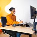 Vrouw met bril en oranje trui schrijft aan een bureau met een documenthouder, naast een computer.