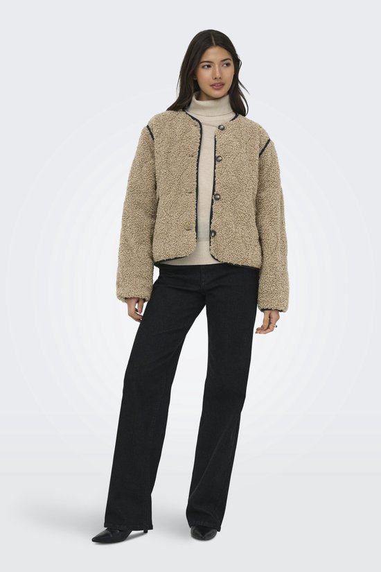 Model draagt een bruine teddy jas met zwarte biezen en knopen, gecombineerd met een coltrui en jeans.