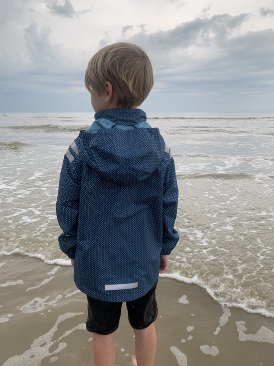 Jongen in een blauw regenpak met visgraatpatroon, staand bij de waterkant op het strand.